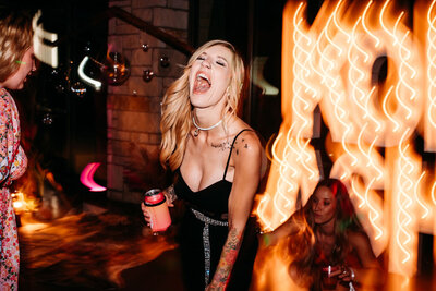 A tattooed alt woman in a black wedding dress is laughing as she dances at her dark and moody reception. The photo is taken with a trendy shutter drag effect causing the marquee lights behind her to streak.