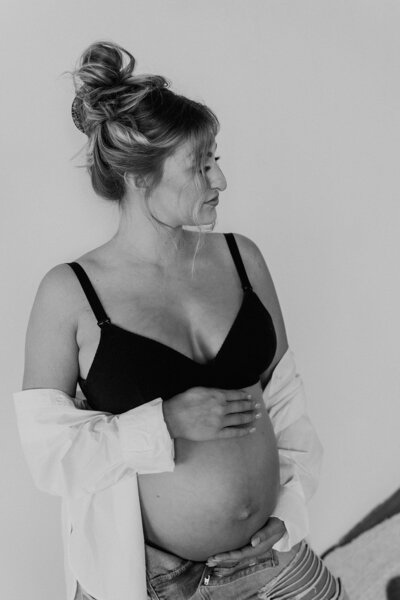 in a classic black-and-white image, a beautiful blonde, pregnant woman wears a black bra and a white button and dress shirt while looking over her left shoulder and holding her pregnant belly.