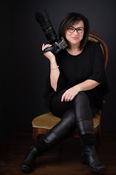 photo of Ottawa Wedding and Portrait Photographer Elenora Luberto JEMMAN Photography dressed in black sitting on a chair holding a camera