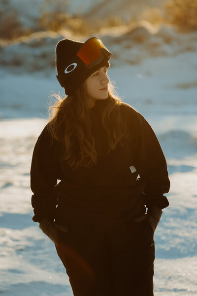 Female destination elopement photographer in ski goggles and winter gear standing in snowy Elnesvågen, Norway, looking into the distance at golden hour.