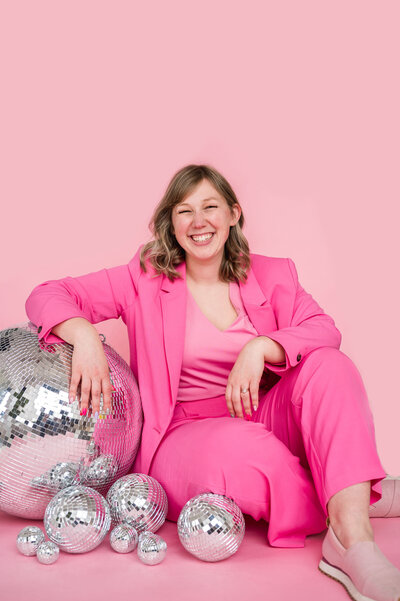 Kat Murphy in a hot pink blazer and pants sitting on the ground next to disco balls in front of a pink background