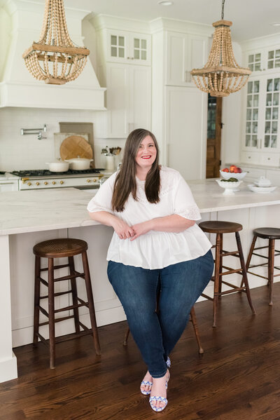 Lauren sits on wooden stool at her kitchen island in white blouse and jeans