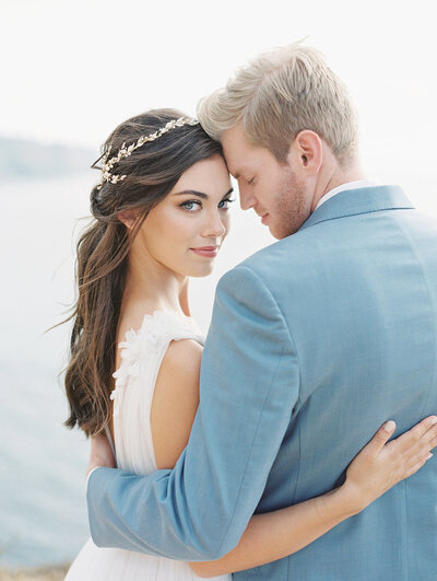Kohnur romantic coastal couple with bride in natural intense makeup and long loose waves with a gold leaf headpiece, hugging near the ocean view.