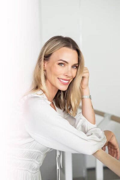 žena s blond vlasy v bílých šatech se opírá o zábradlí a dívá se s úsměvem do objektivu