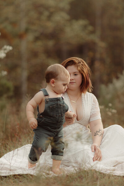 Mama in white dress holds hands with baby boy in denim overalls by Lawrenceville Georgia Baby Photographer