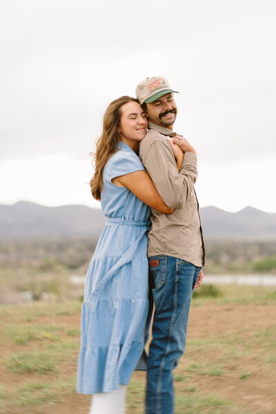 Woman hugging fiancé from behind, smiling at camera