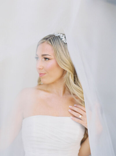 Wedding Photographer RI | A bride in a strapless gown with long blonde hair and a jeweled headpiece stands behind a sheer white curtain, gazing thoughtfully to the side.
