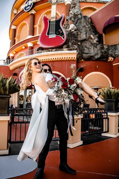 Shannon Lee, a Florida elopement photographer capturing a fun and playful moment of a bride being swept off her feet by her partner outside a vibrant venue with a giant guitar.