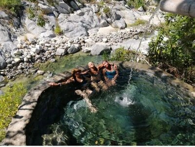Three women soaking in a circular stone hot spring pool surrounded by river rocks and lush canyon greenery, enjoying a refreshing water stream and tranquil mountain energy at Corazón del Agua.