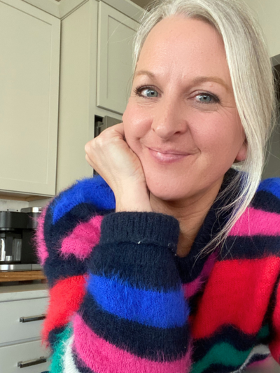 Woman with blonde hair wearing a colorful striped sweater, smiling while sitting in a kitchen.