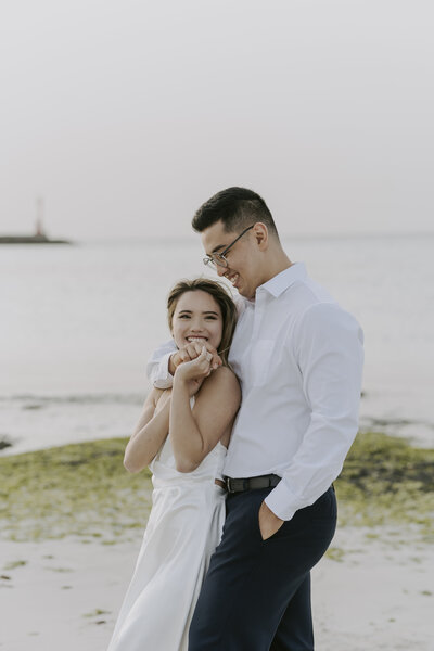 woman in white silk dress smiles with her husband puts his arm around her