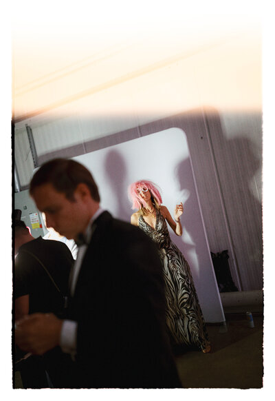 April M Payne Photography captured a candid moment at a wedding reception, with a guest in a bold outfit striking a pose in front of a backdrop, while a man in a tuxedo walks past, partially in the frame.
