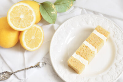 Slice of Sicilian lemon and elderflower cake on a white plate with lemons on a linen tablecloth