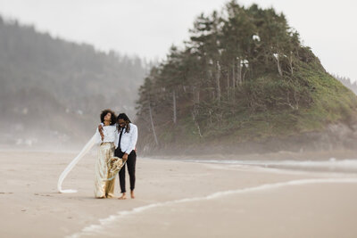 A BIPOC bride wearing a shimmering gold skirt , white cropped blouse and white long veil blowing in the wind, walks hip to hip with her dreadlocked Black groom wearing black slacks barefoot along the beach at their Oregon Coast wedding and elopement near Lincoln City. | Erica Swantek Photography