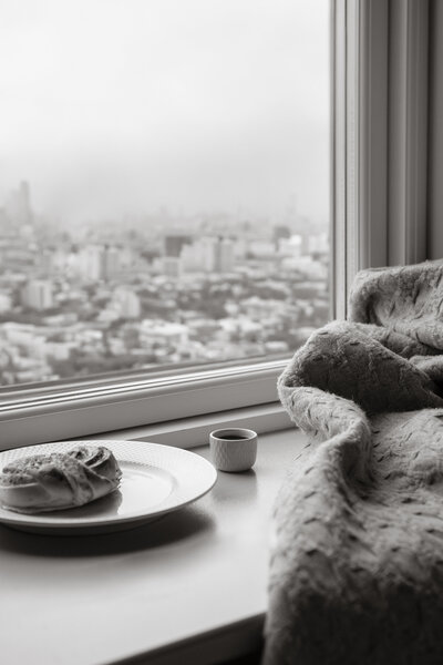 Black and white photograph of a cozy breakfast scene on a windowsill overlooking a cityscape. A knitted blanket drapes beside a small cup of coffee and a white plate holding a pastry. The out-of-focus skyline appears under a hazy sky.