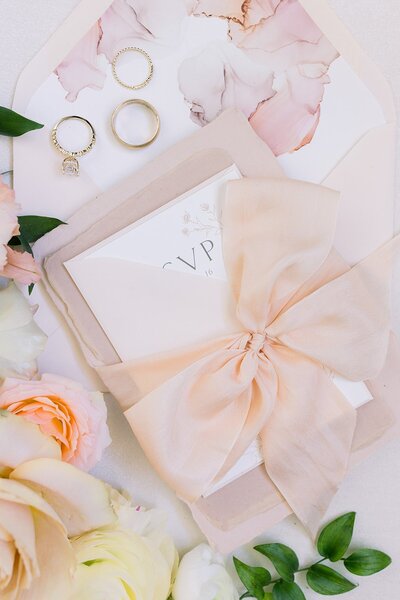 Elegant wedding invitation suite wrapped in a silk bow, styled with gold rings, floral accents, and soft textures. Designed by a Central Coast wedding planner.