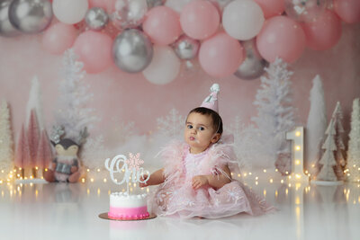 1st Birthday girl wearing pink snowflake dress with party hat on with her smash cake on a pink winter wonderland background