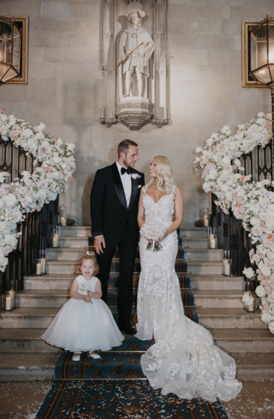 Danielle Armstrong and Tommy Edney on their UK wedding day. Danielle in a Berta dress, Tommy in a tuxedo, framed by a green archway.
