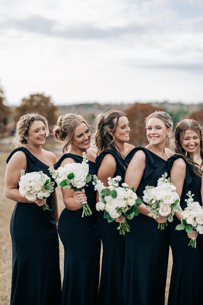 women in black dresses smiling and looking at each other while holding white bouquets