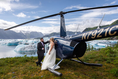 Bride and groom stand beside a helicopter on a remote Alaskan glacier lake, surrounded by floating icebergs.