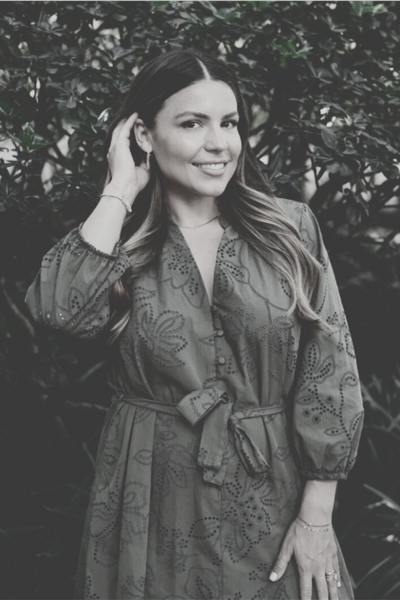 Natalie Bernacchi standing against a light-colored textured wall, smiling softly, wearing a belted olive green eyelet dress with gold jewelry and long wavy hair offering real-time mindset, business, and energetic support through private messaging and voice notes.