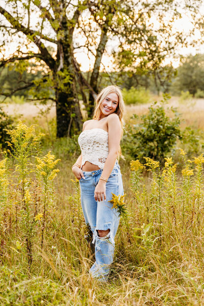 stunning young lady in a white lace top and ripped jeans walking through a beautiful field at sunset captured by Ashley Kalbus Photography