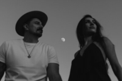 A black and white photo captures a man in a hat and a woman, their gazes diverging, as the moon hangs in the clear sky between them—a timeless moment through the lens of a Toronto wedding photographer.