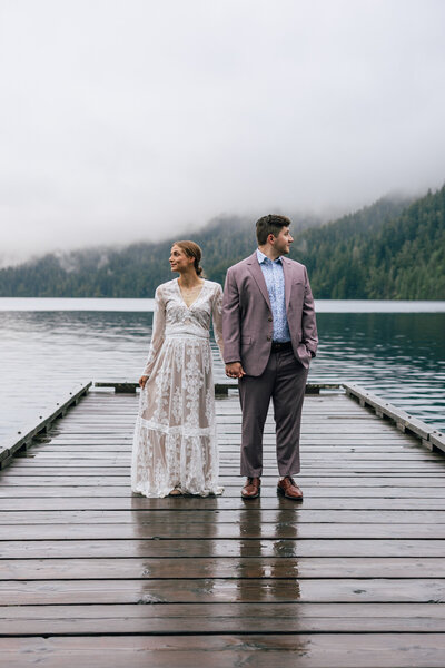 A groom in a mauve suit holds the hand of his bride in a white lace gown standing on the dock of Lake Crescent for their Olympic National Park Elopement in Washington State.