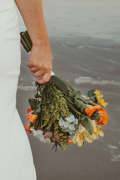 BRIDE HOLDS COLORFUL BOUQUET TO HER SIDE WITH  SAND FROM THE PORT ARANSAS TEXAS BEACH IN THE BACKGROUND