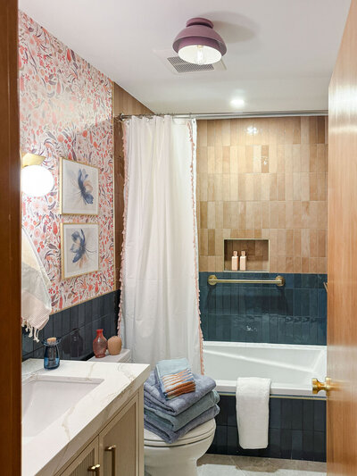 Full bathroom with pink and emerald tiled bath tub, white vanity and toilet, and floral wallpaper