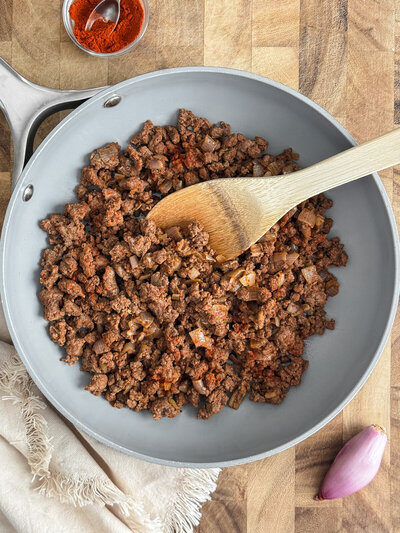A pan of ground bison meat with chopped shallots, a wooden spoon, with a bowl of paprika nearby.