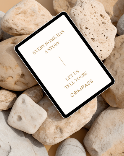 A tablet displaying a Compass real estate ad with the text, "Every home has a story. Let us tell yours," placed on a pile of light-colored rocks, highlights thoughtful brand design by Sweet Pea Ink Creative.