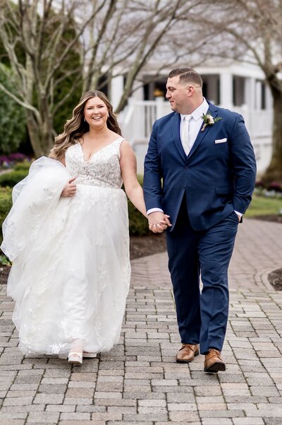 A bride in a white long flowing gown holding hands with her groom who is wearing a navy suit and brown shoes.  They are walking towards the camera.  The groom looks at his bride adoringly as she smiles at the camera outside their wedding venue The Mill Lakeside Manor in NJ.