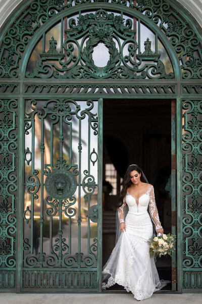 A bride in an elegant gown stands behind a beautifully ornate iron gate, embodying the timeless elegance and sophistication of a luxury wedding setting.