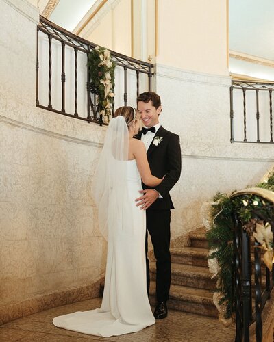 A bride and groom stand on a staircase inside a building, smiling and looking at each other. The bride wears a white dress and veil, while the groom sports a black tuxedo. Thanks to their wedding planner in Banff, every detail was perfect for their special day.