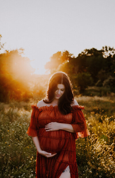 A mom in a rust color maternity gown standing in a grassy field looking down at her belly at Northwest Community Park in Fort Worth, Texas