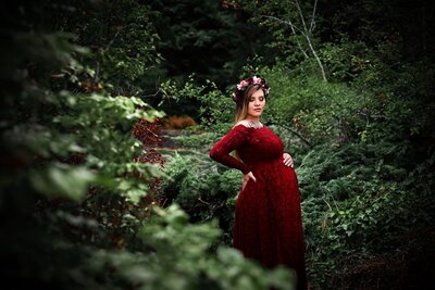Pregnant woman wearing a long sleeve red lace dress during outdoor maternity session at Whytecliff Park in North Vancouver surrounded by greenery and trees.