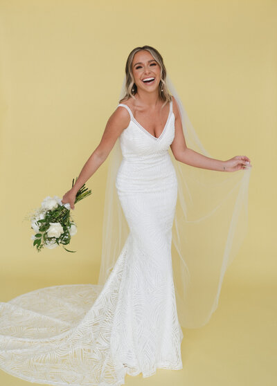 Bride holding her veil and bouquet and smiling in front of a yellow studio background
