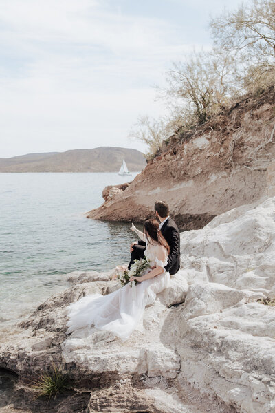Bride and groom sitting on white rocks pointing out at a sailboat going by at Lake Pleasant during their Arizona elopement.