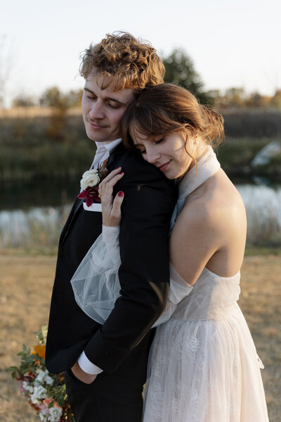 Loving eloping couple stands together in a quiet field with water in the background, wrapped in love and nature. 
