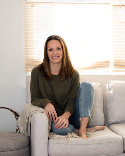 kelly francis siting on neutral sofa with natural sunlight green sweater and light denim jeans
