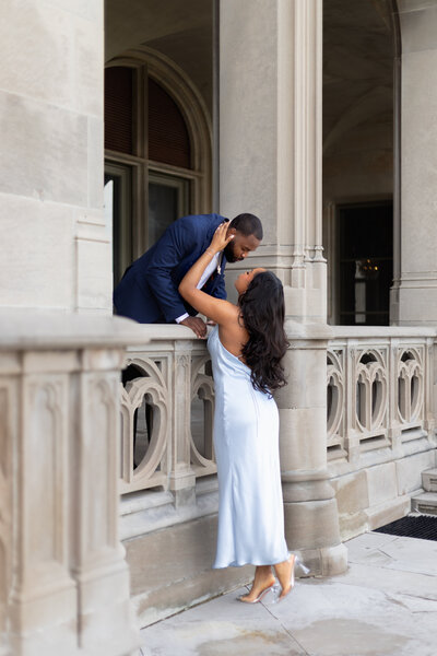Rhode Island Wedding Photographer | Kelsey Sheehan Photography Timeless Rhode Island Weddings | A woman in a white dress and a man in a navy suit share a romantic moment on an ornate stone balcony, conveying love and intimacy.