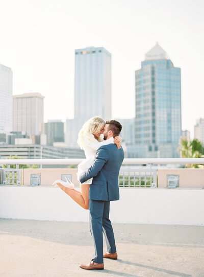 engagement session in Downtown Tampa featuring the Tampa skyline Groom lifting bride up both smiling Tampa engagement session location