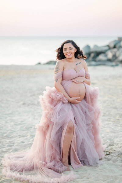 An expectant mother poses on the beach in a pink tulle gown. She is all smiles while holding her belly.