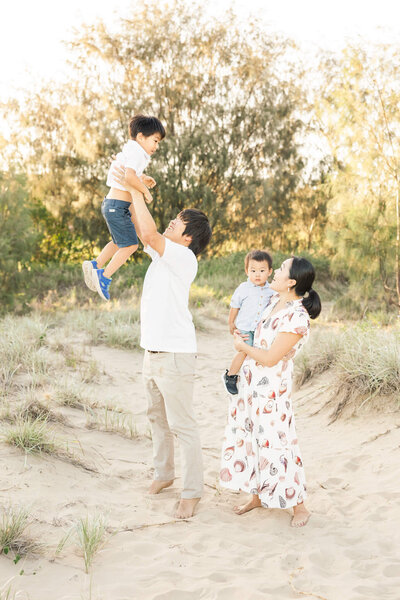 Family portrait of dad and mum with two boys, dad is throwing one boy in the air while mum holds other little boy and looks on.