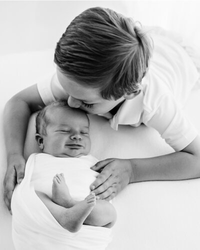 a big brother kisses his newborn sibling on the head in a classic black and white photo