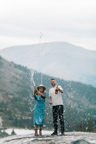 engaged couple spraying champagne on top of a mountain with hat on during engagement session