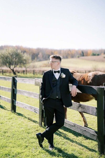Groom in black tuxedo leaning on wooden fence with horse behind, stylish wedding portrait in Ontario countryside
