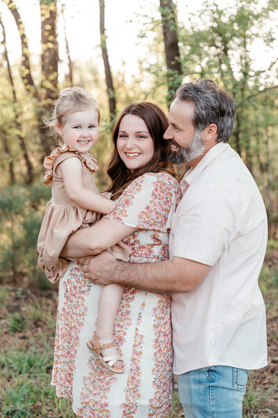 "Smiling family standing outdoors in a sunlit forest, featuring a mother in a floral dress, a father in a white shirt, and their young daughter in a beige dress with ruffled sleeves. A warm and joyful family photography moment."