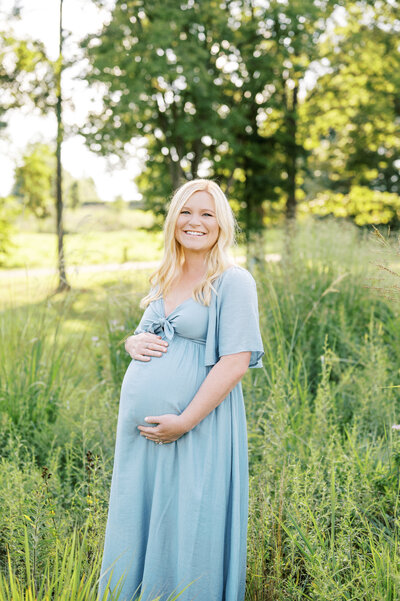 Mom holds belly and smiles at the camera in field of tall grass during Summer maternity session